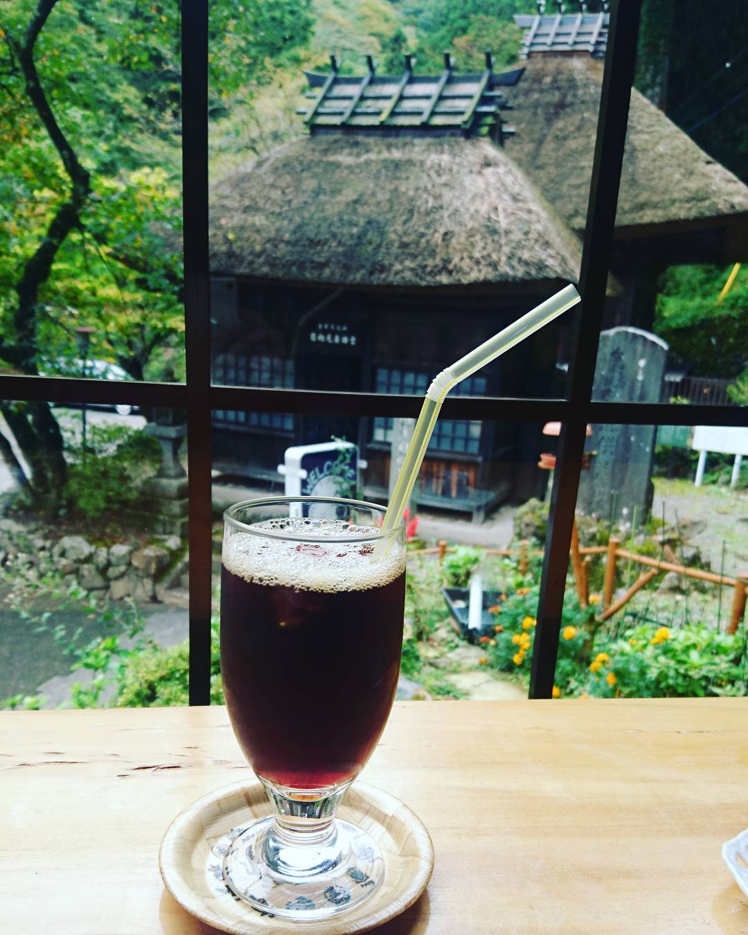 かやぶき屋根の建物を眺めるカフェのアイスコーヒー