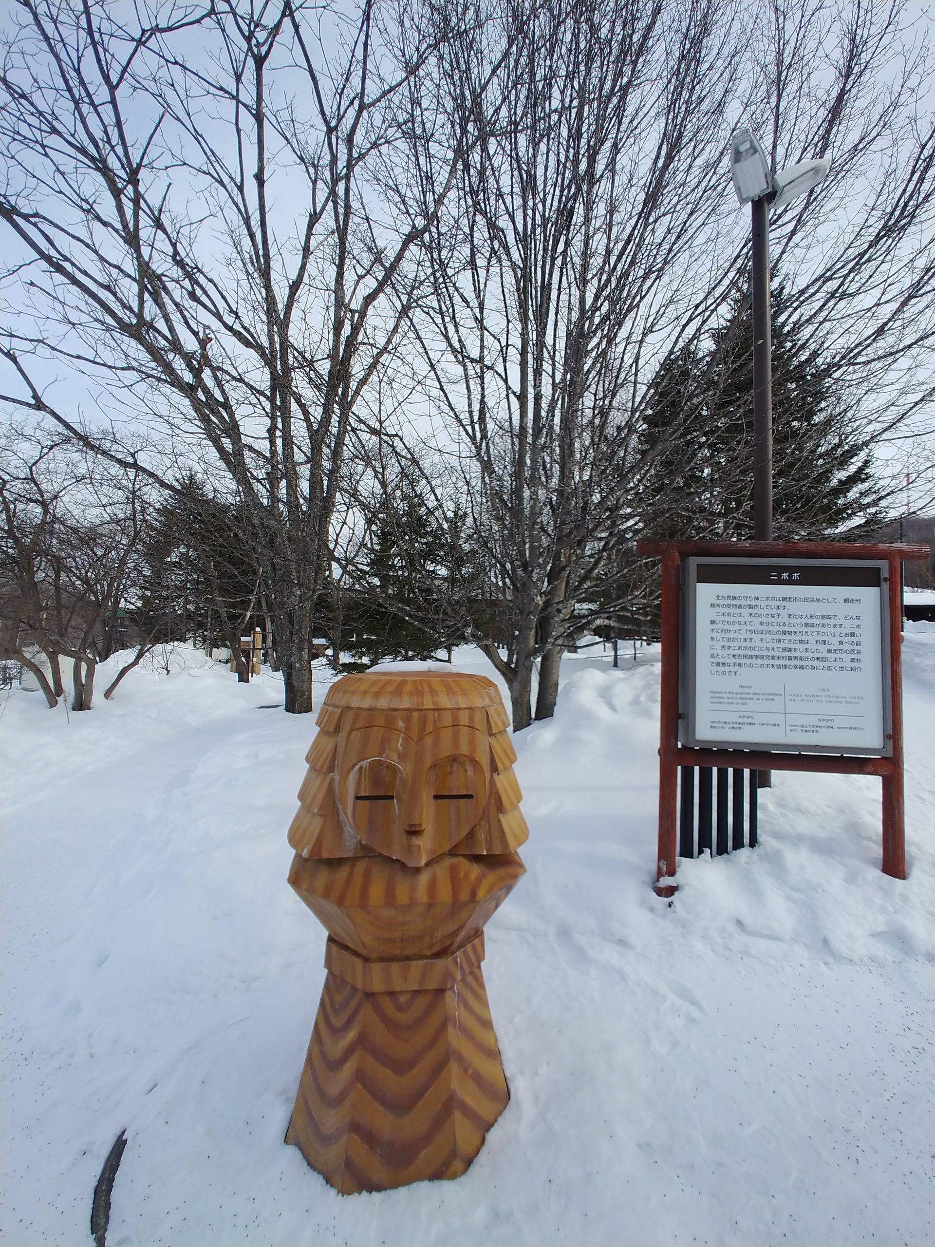 雪の中に立つ木彫りの像と説明板