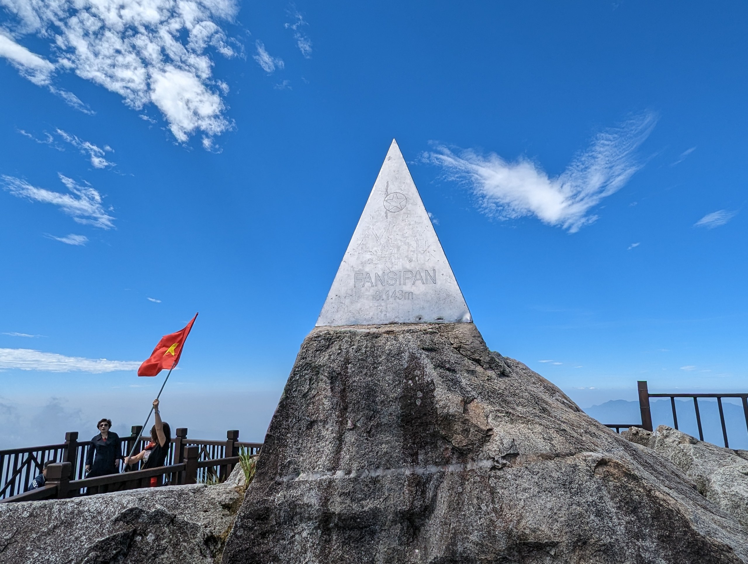国旗がはためく岩の上の、銀色の三角の山頂標