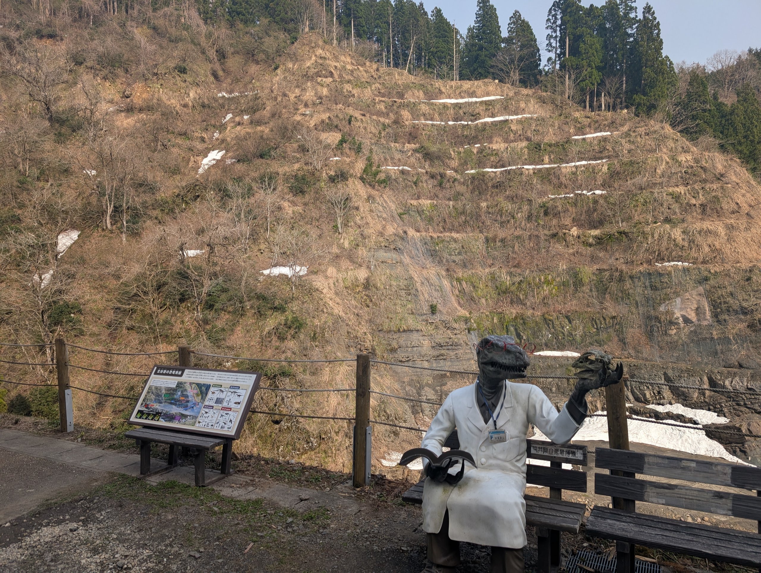 しま模様の地層が見える崖と、ベンチに座る人の像と案内板