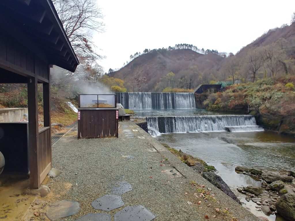湯気が上がる川沿いの道と、段になった堰のある肘折温泉の風景