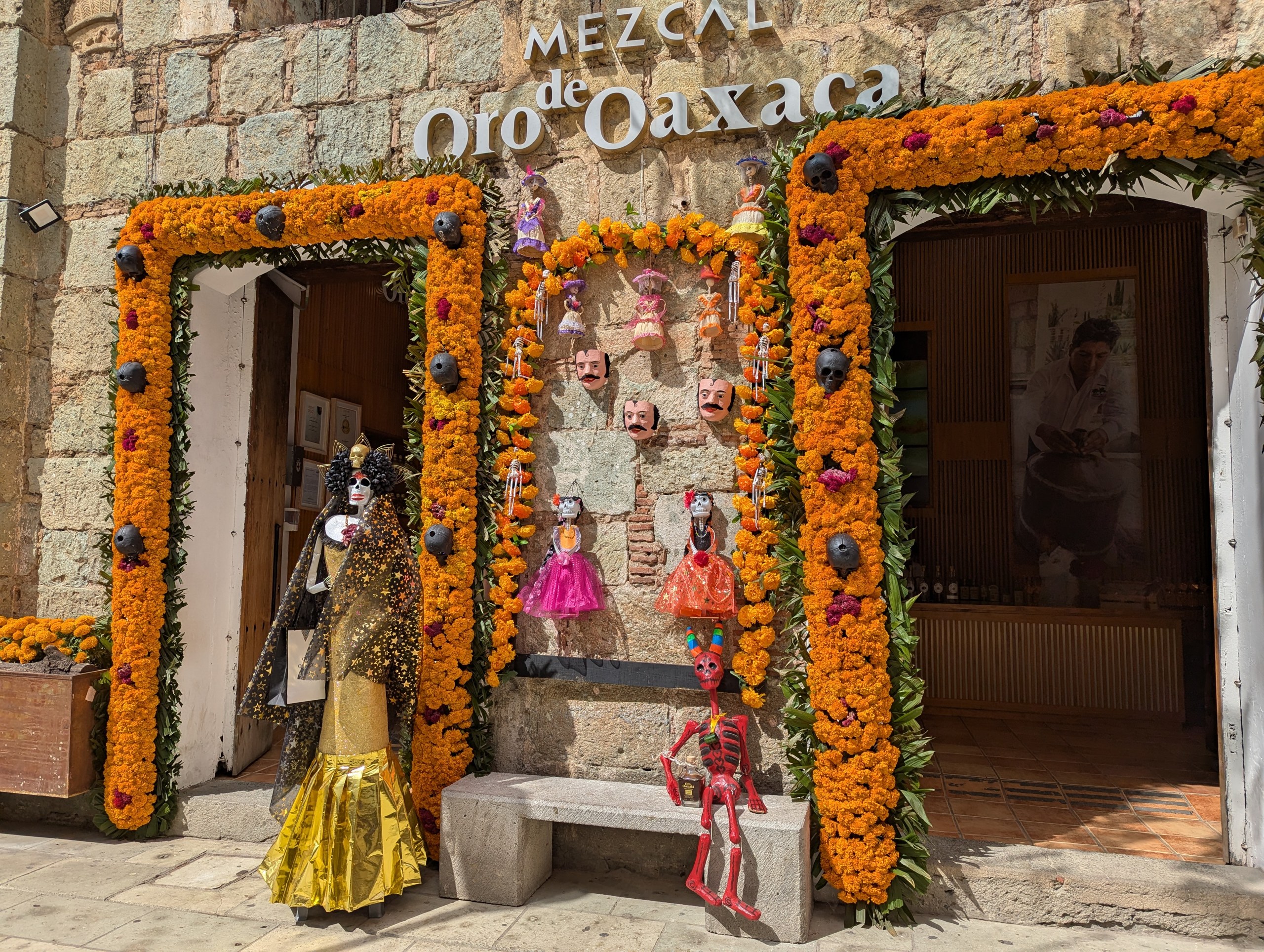 「Mezcal Oro de Oaxaca」の看板の下、マリーゴールドで縁取られた入口とガイコツの人形