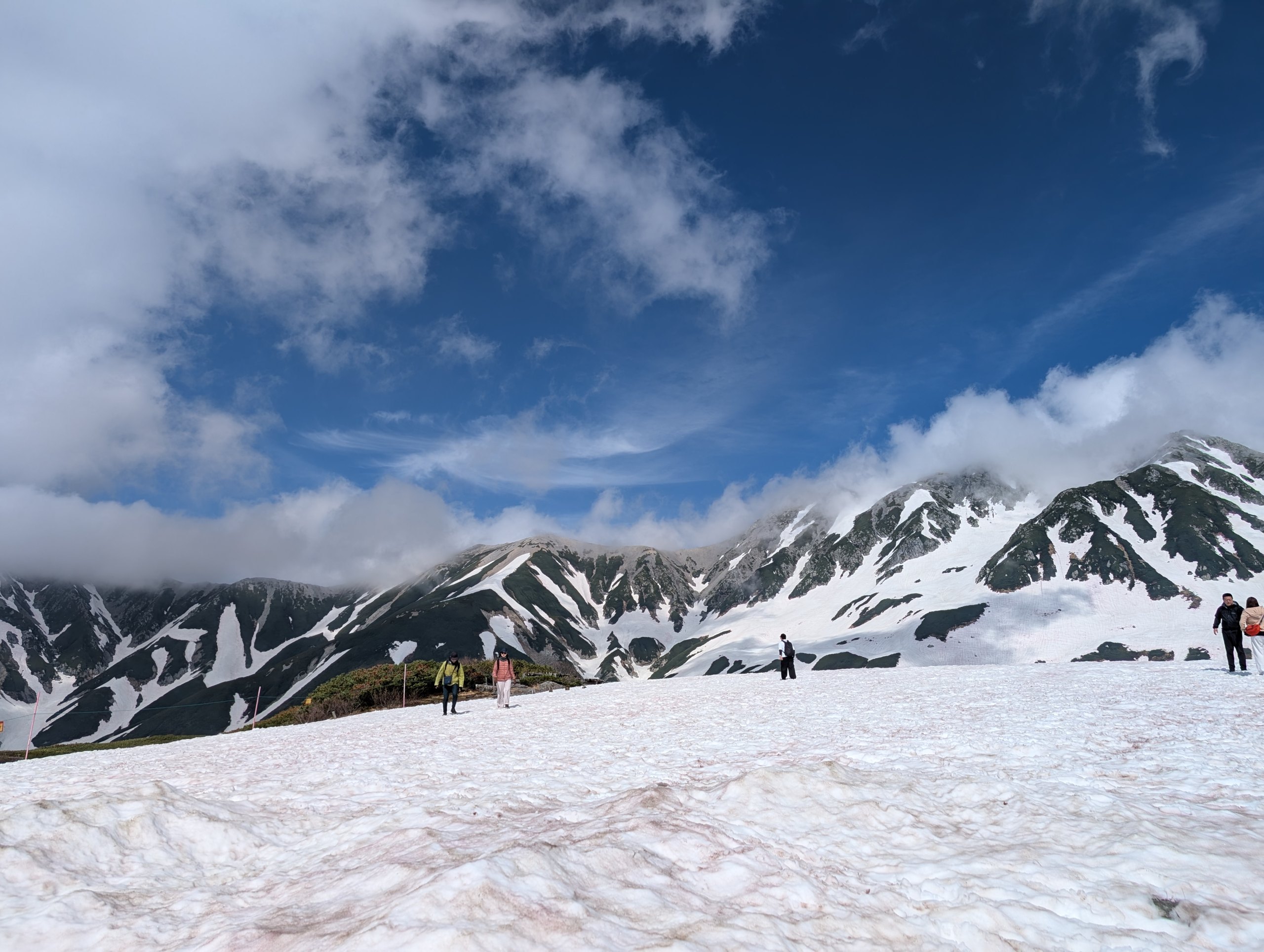 雪の上を歩く人と、雪の残る山並み
