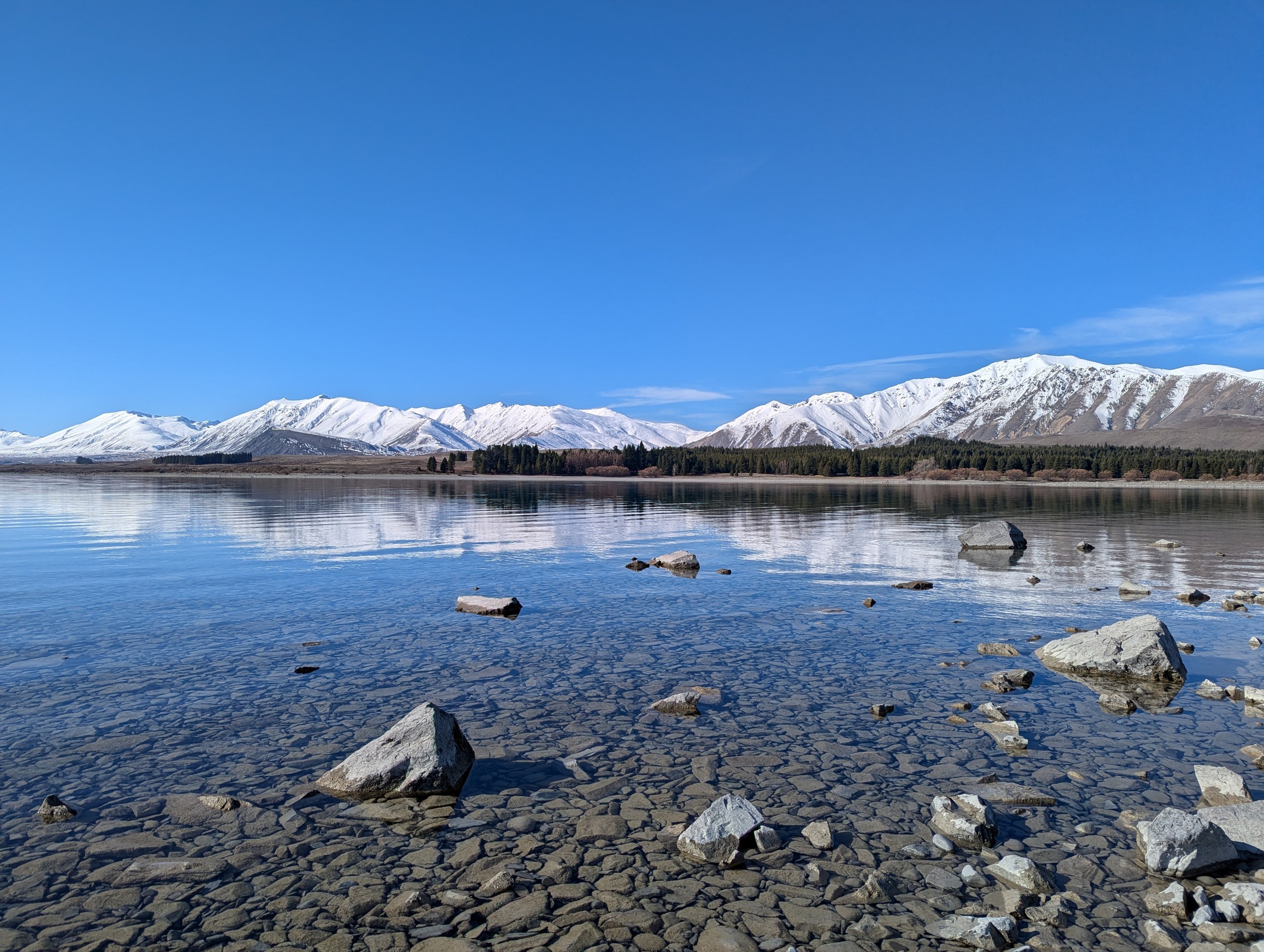澄んだ水に石が透けて見える湖岸と、湖の向こうの雪山