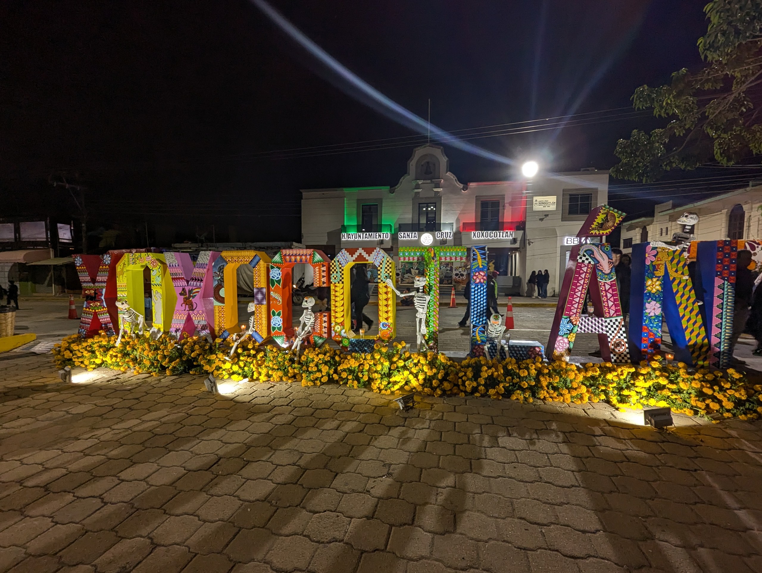 夜の広場に置かれた「XOXOCOTLAN」のカラフルな大きな文字と、足元のマリーゴールド