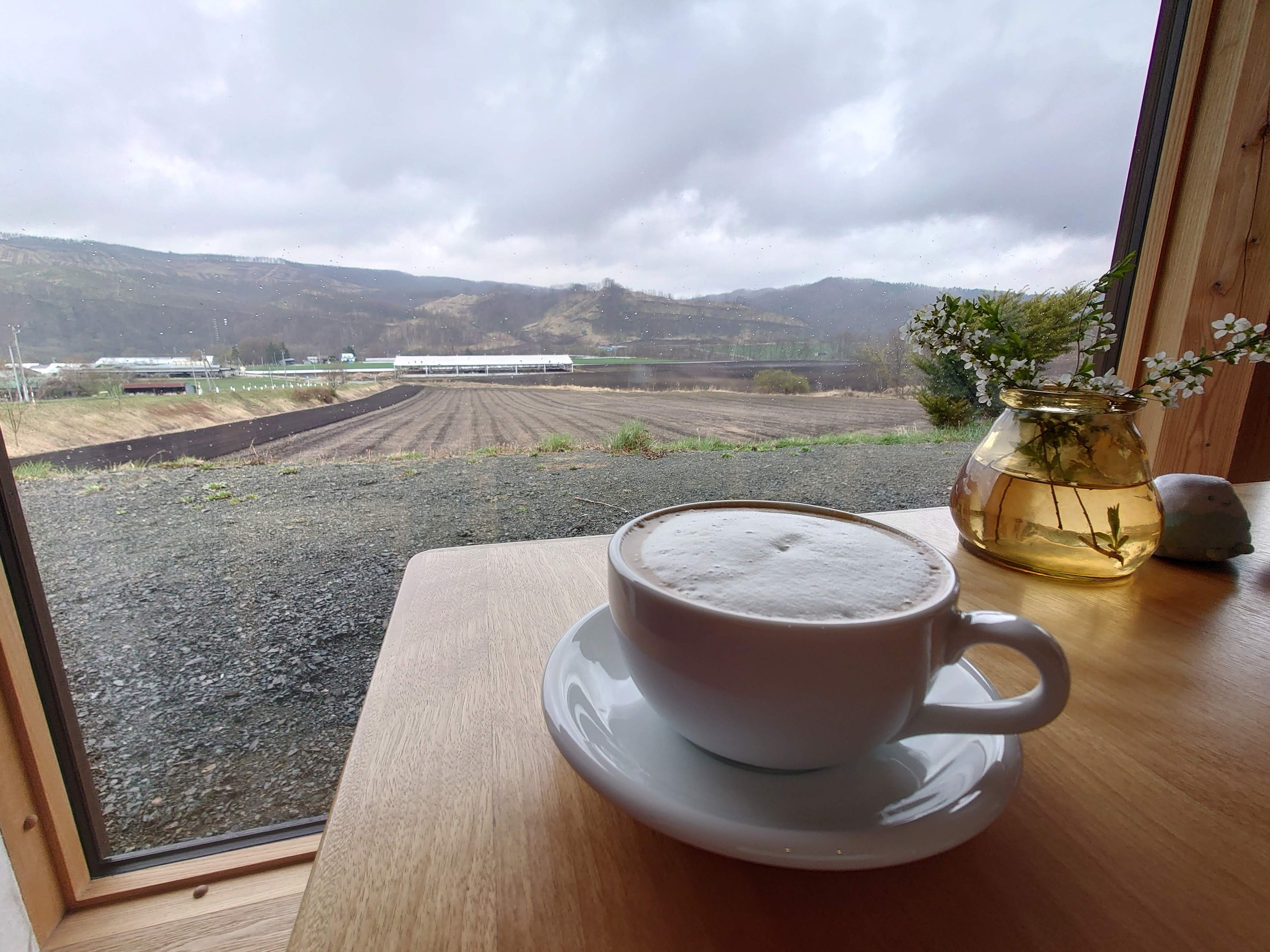 窓辺のカフェラテと、外に広がる畑と山並み