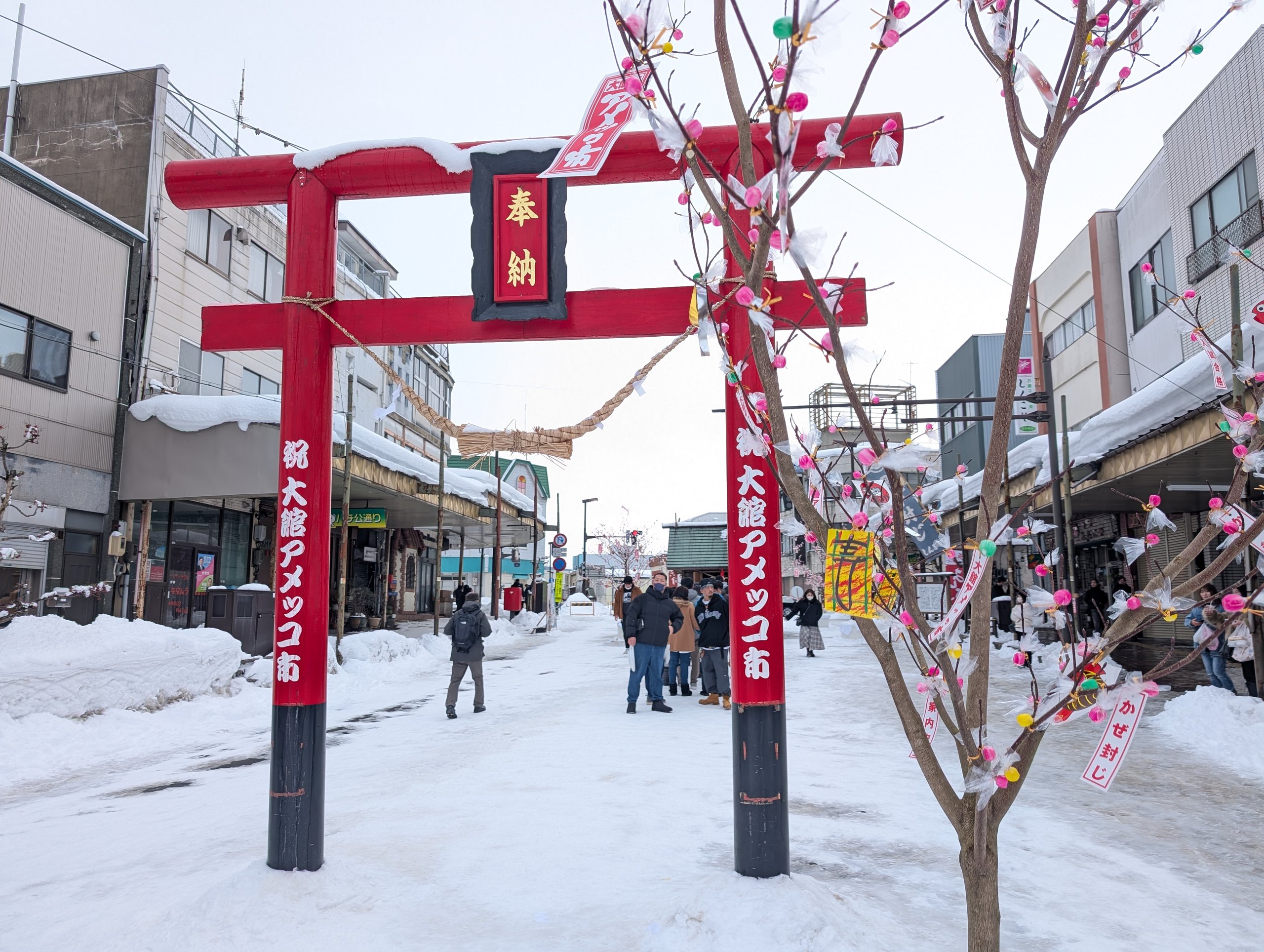 「大館アメッコ市」と書かれた赤い鳥居と、雪の通り