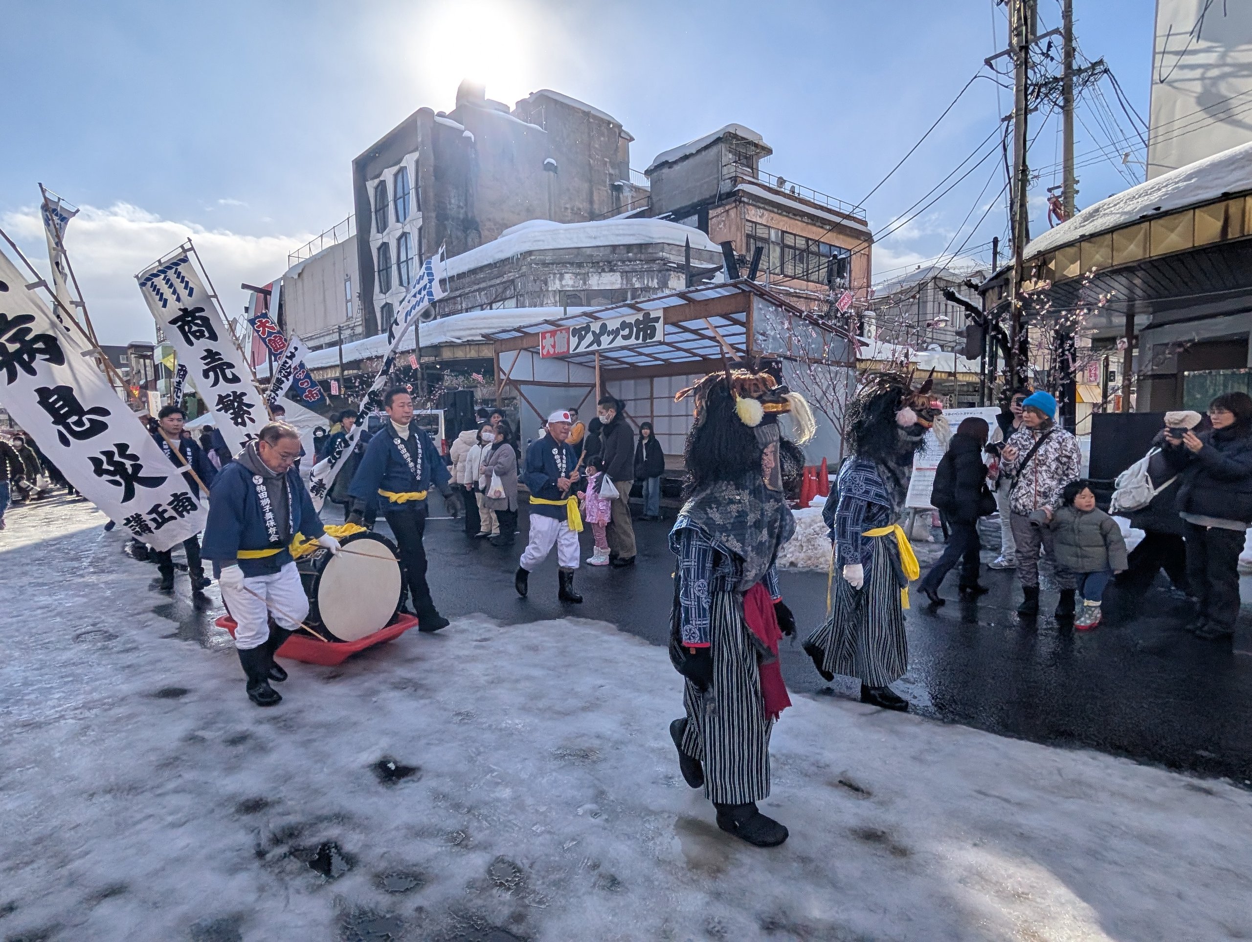 のぼりを持った人や太鼓を引く人が、雪の通りを進む行列