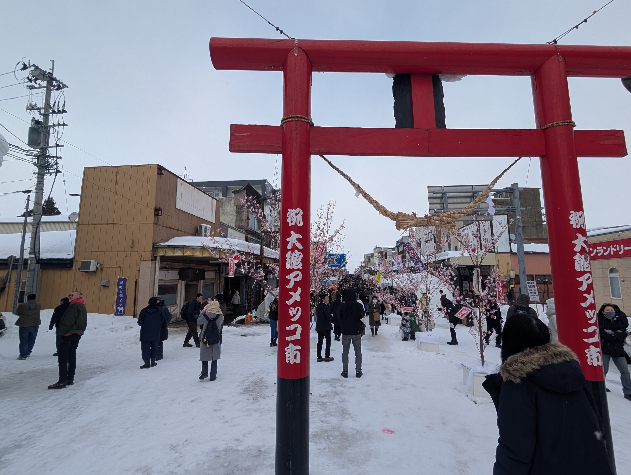 「大館アメッコ市」と書かれた赤い鳥居と、雪の通りを歩く人たち