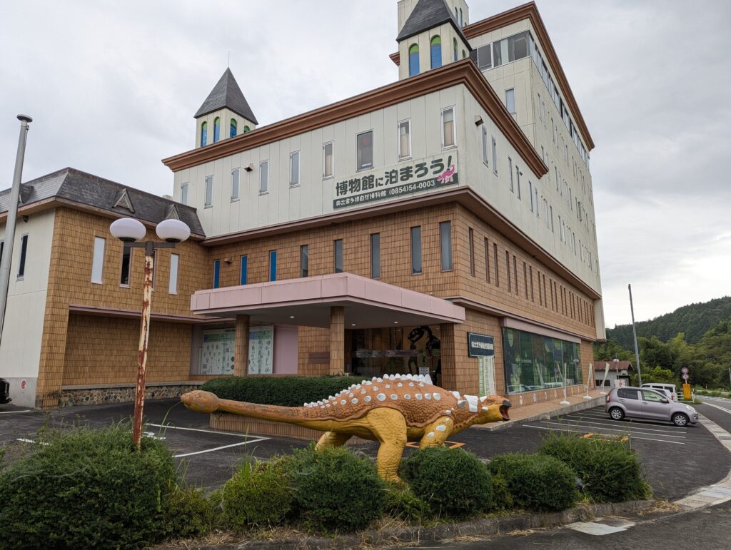 「博物館に泊まろう！」の看板がかかる建物と、入口前のオレンジ色の恐竜の像