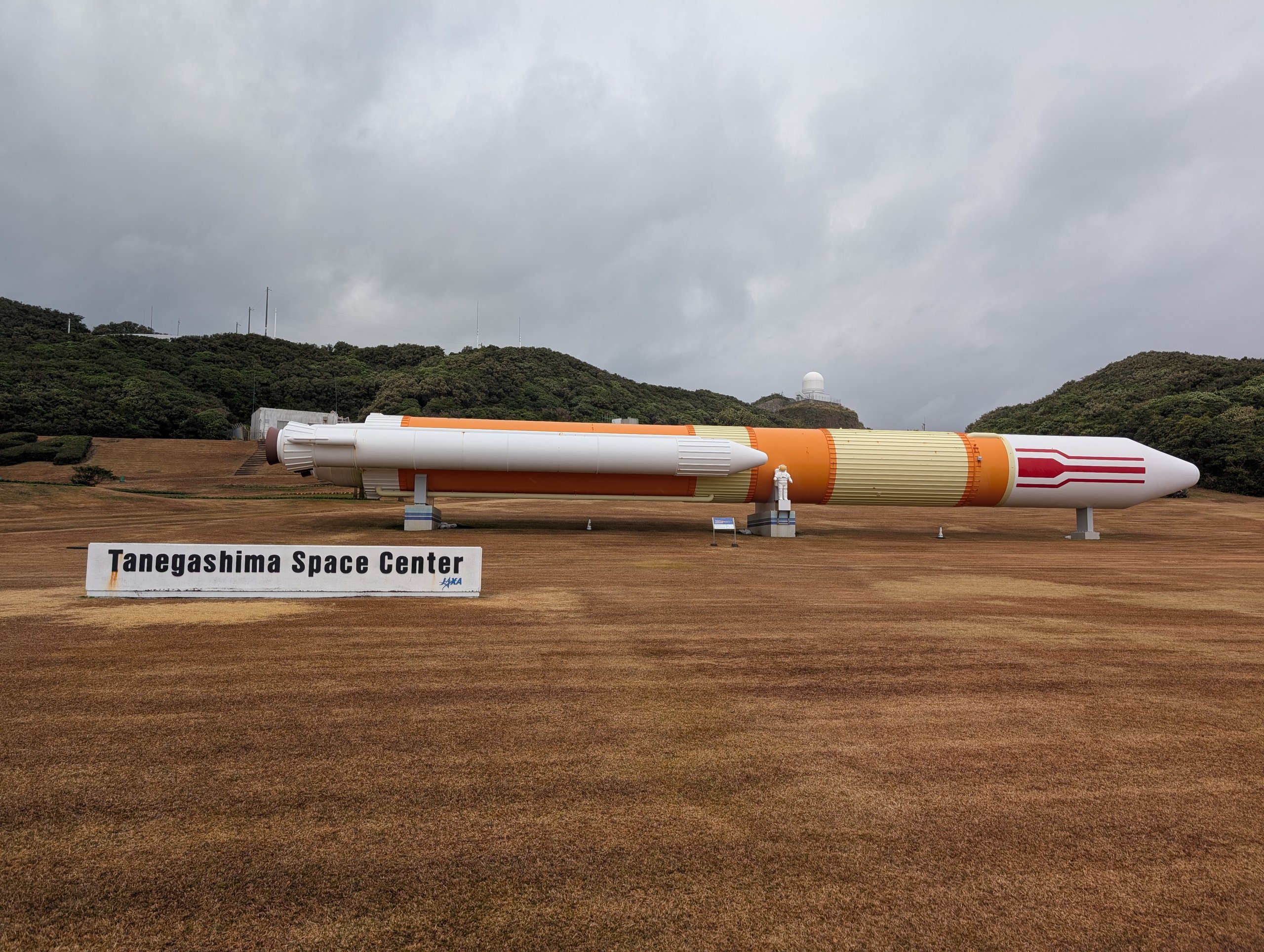 芝生の上に横向きに置かれた白とオレンジの大きなロケットと、「Tanegashima Space Center」の看板