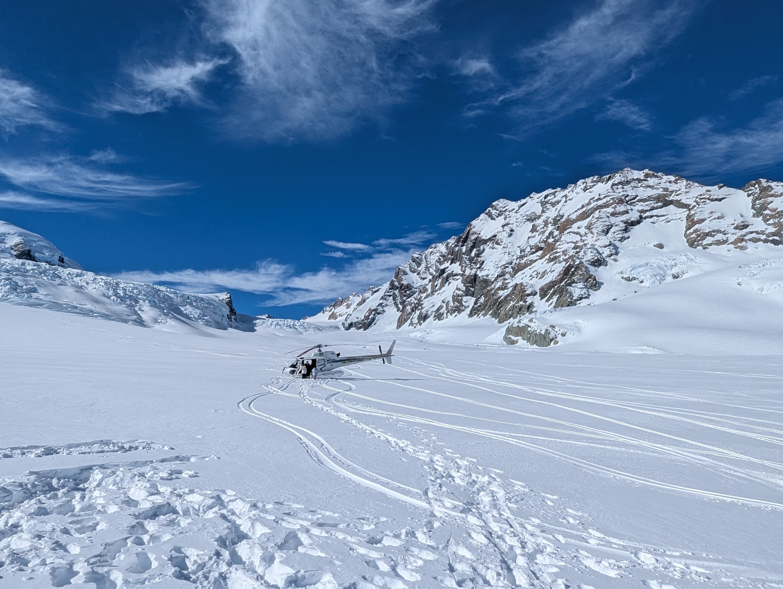 真っ白な雪原にぽつんと止まるヘリと、その奥の岩肌の山