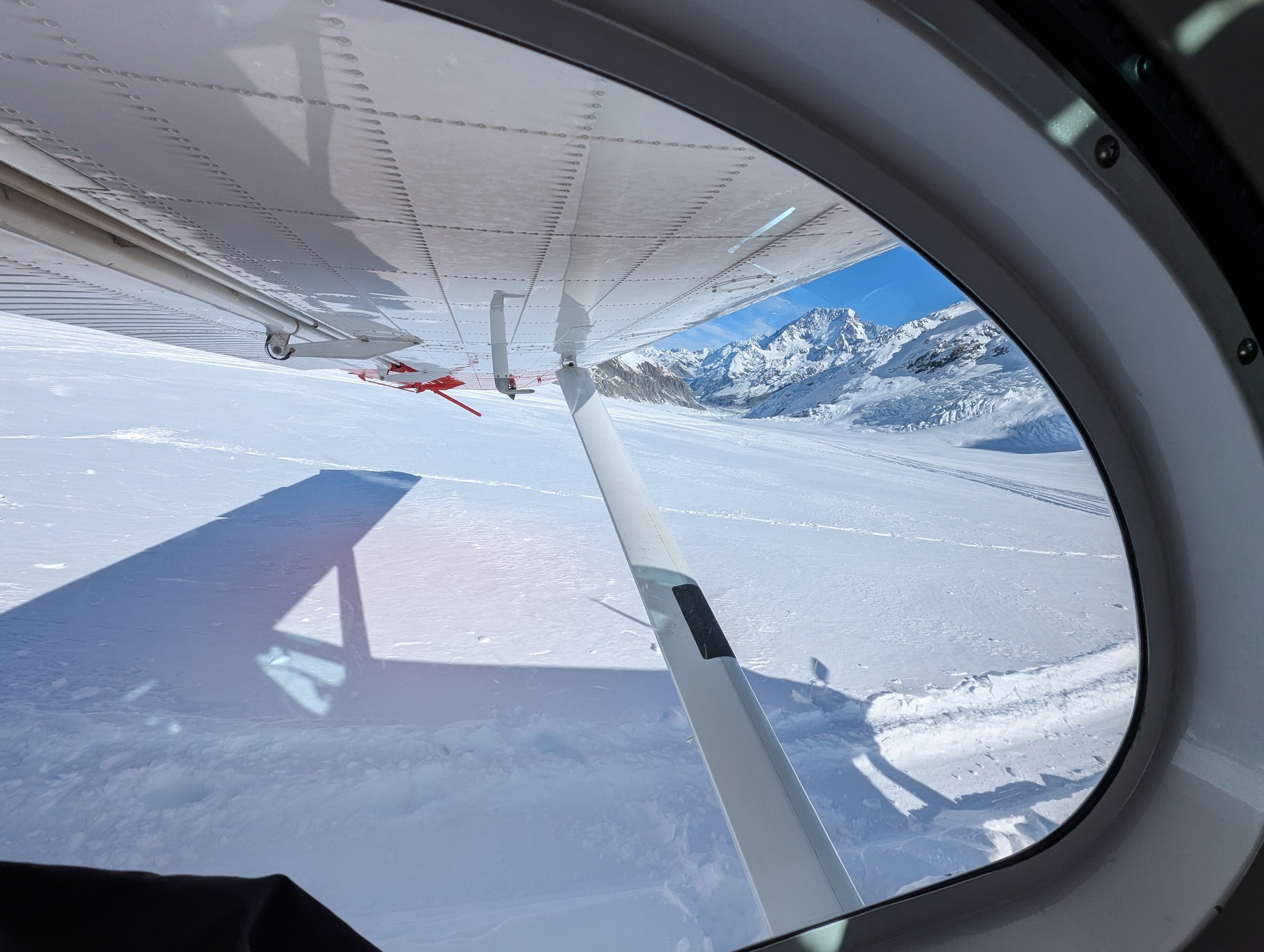 スキープレーンの窓から見た、翼と支柱、その下に広がる雪の氷河と山