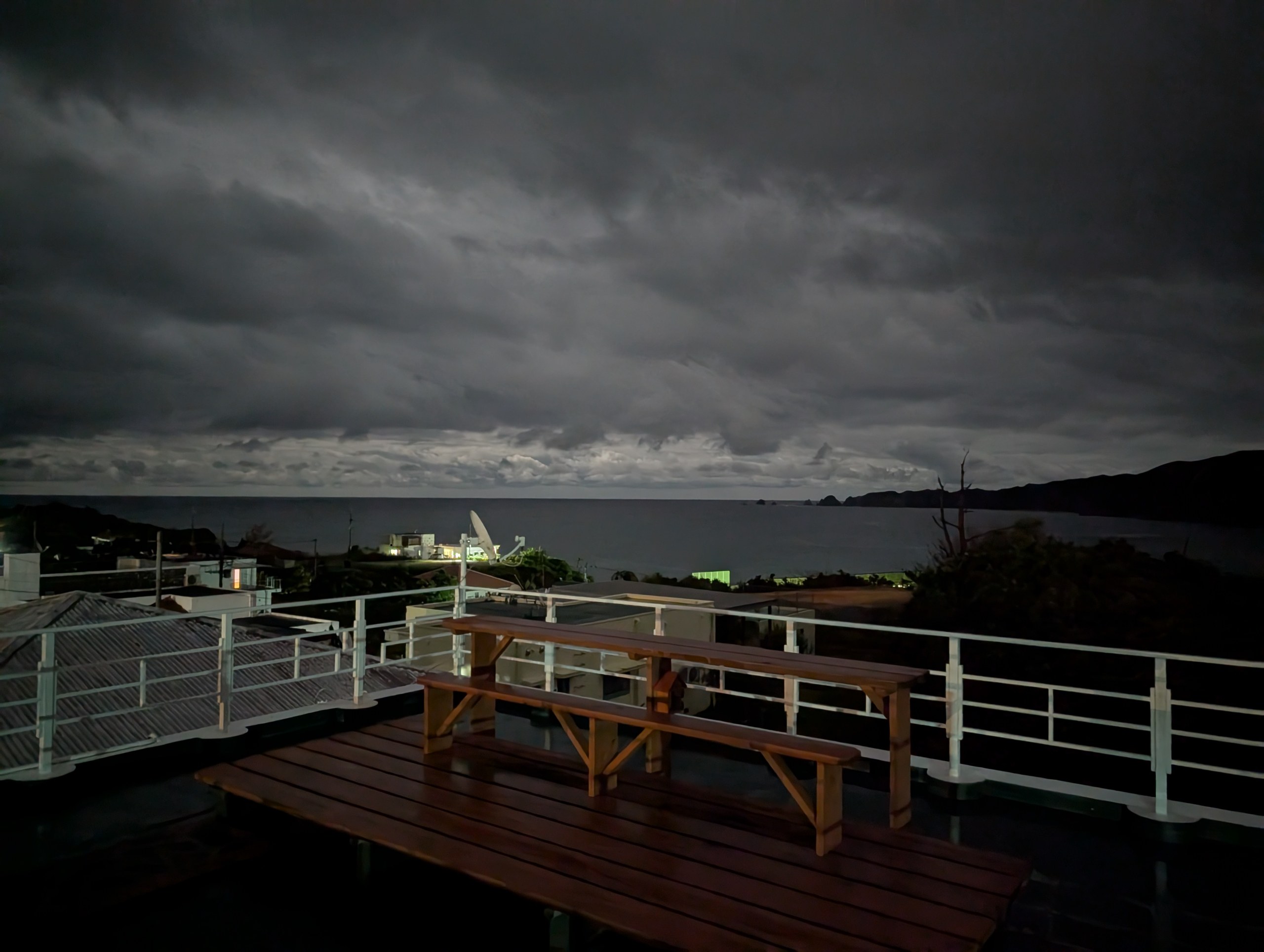 夜の芭蕉の屋上。木のテーブルとベンチの向こうに、厚い雲におおわれた空と海