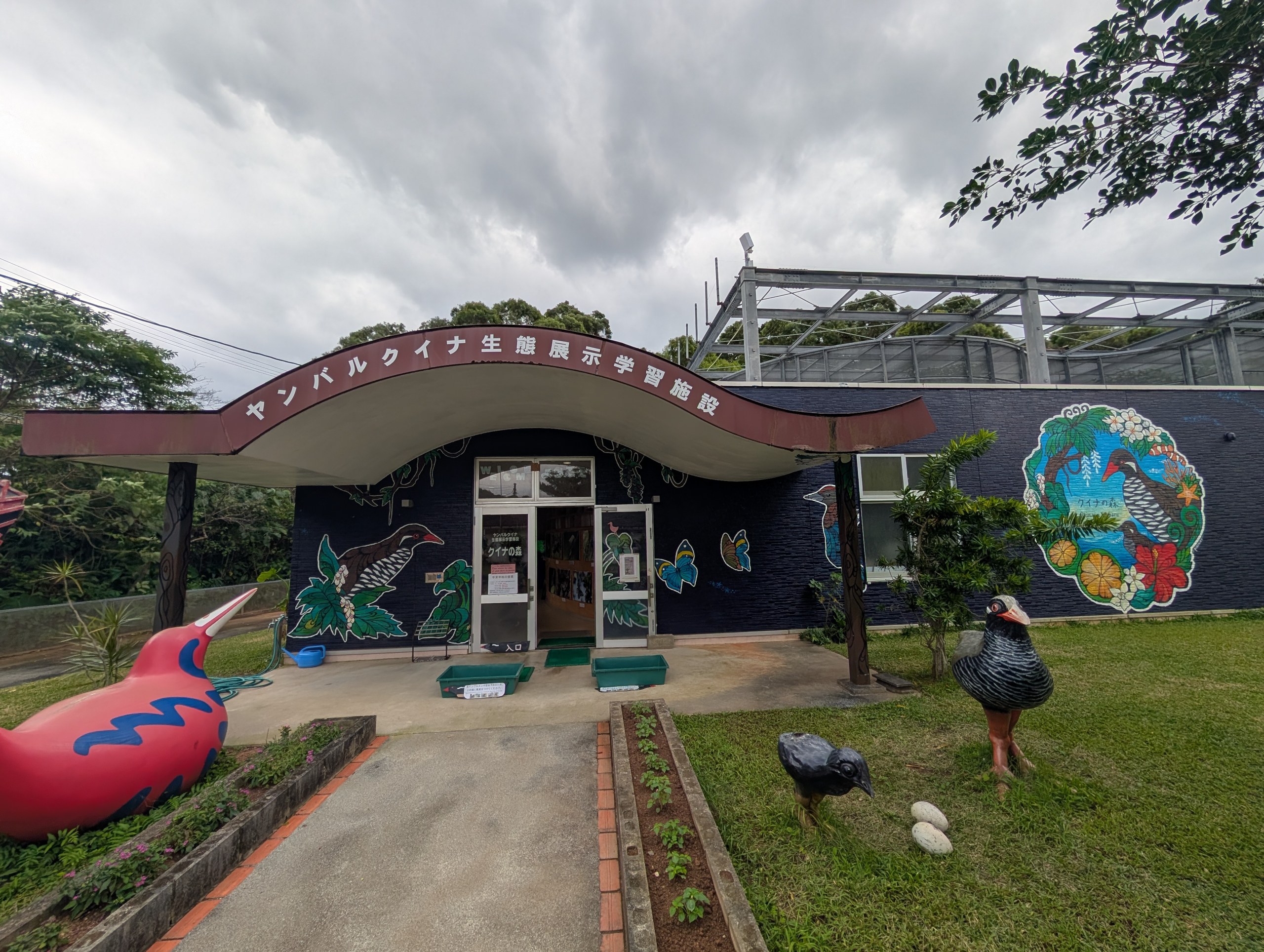 「ヤンバルクイナ生態展示学習施設」の看板の建物。壁にヤンバルクイナの絵、前庭に赤い鳥の像と黒いヤンバルクイナの像