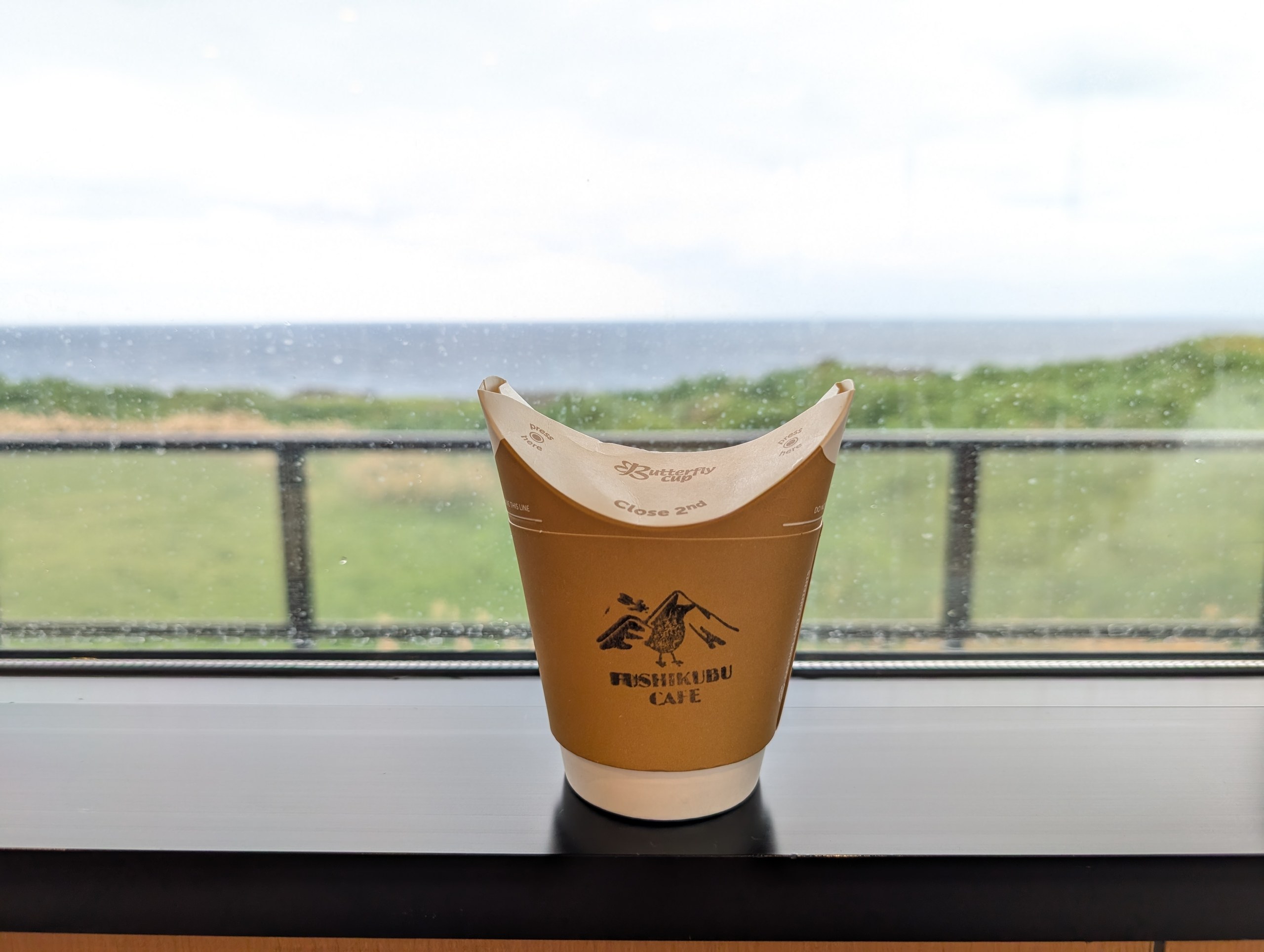 窓辺に置かれた「FUSHIKUBU CAFE」の紙カップ。窓の外に草地と海