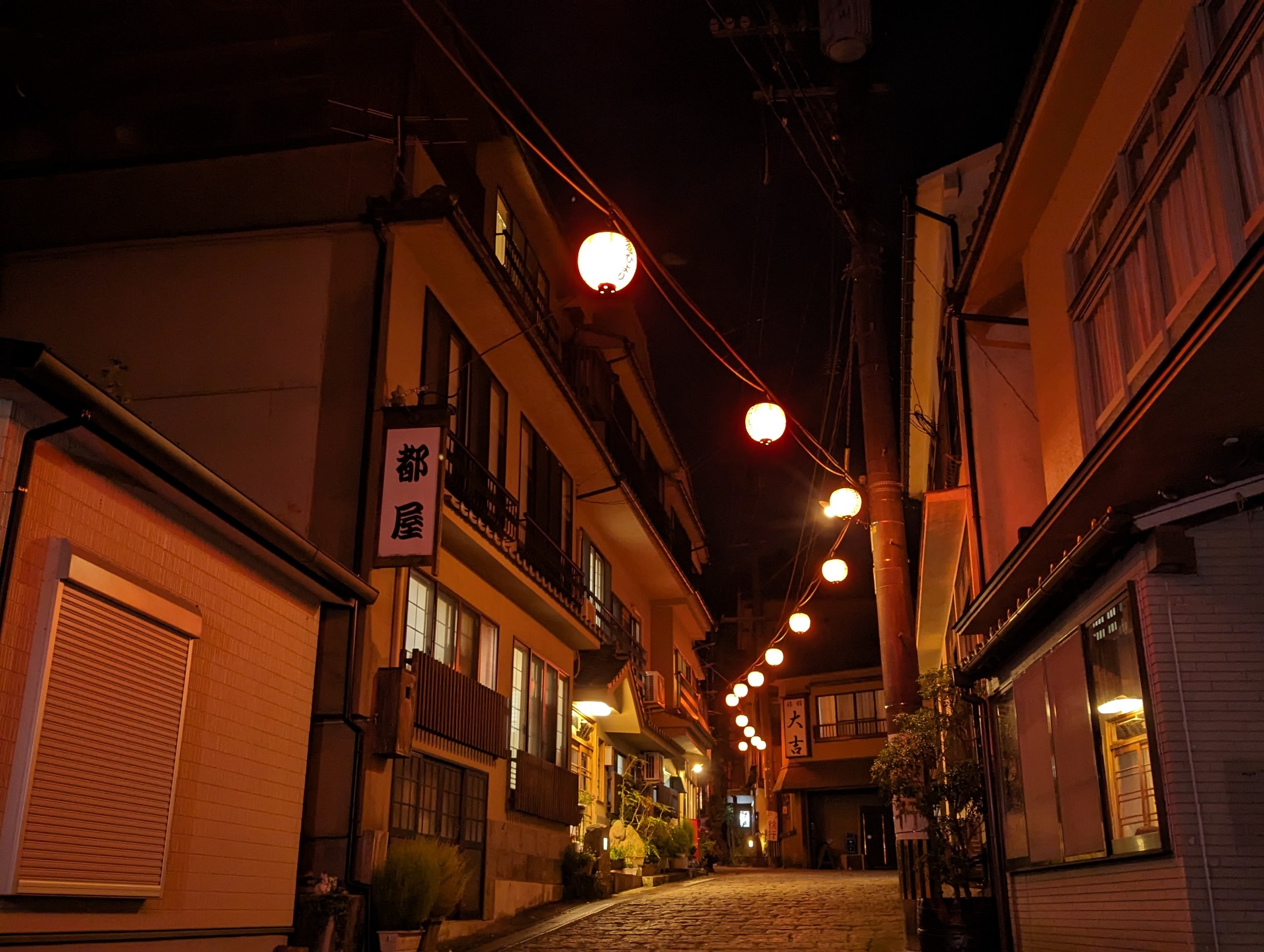 「都屋」の看板と、提灯が続く夜の石畳の通り